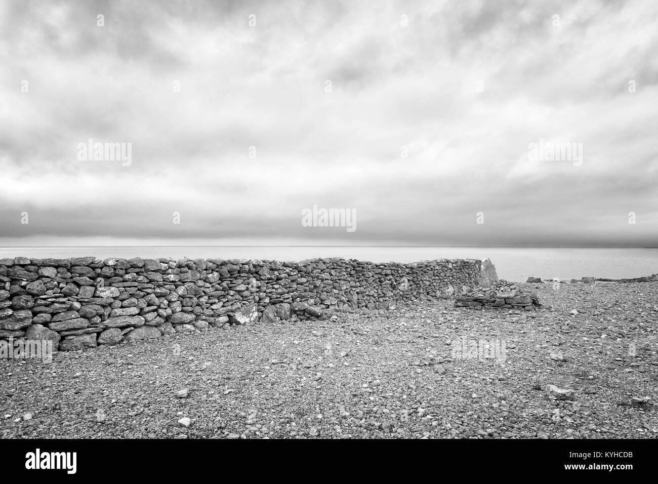 Old stone wall on a rocky beach runs perpendicular to the edge of the sea. Moody mysterious scene. Heavy clouds. Black and white. Faro, Sweden Stock Photo