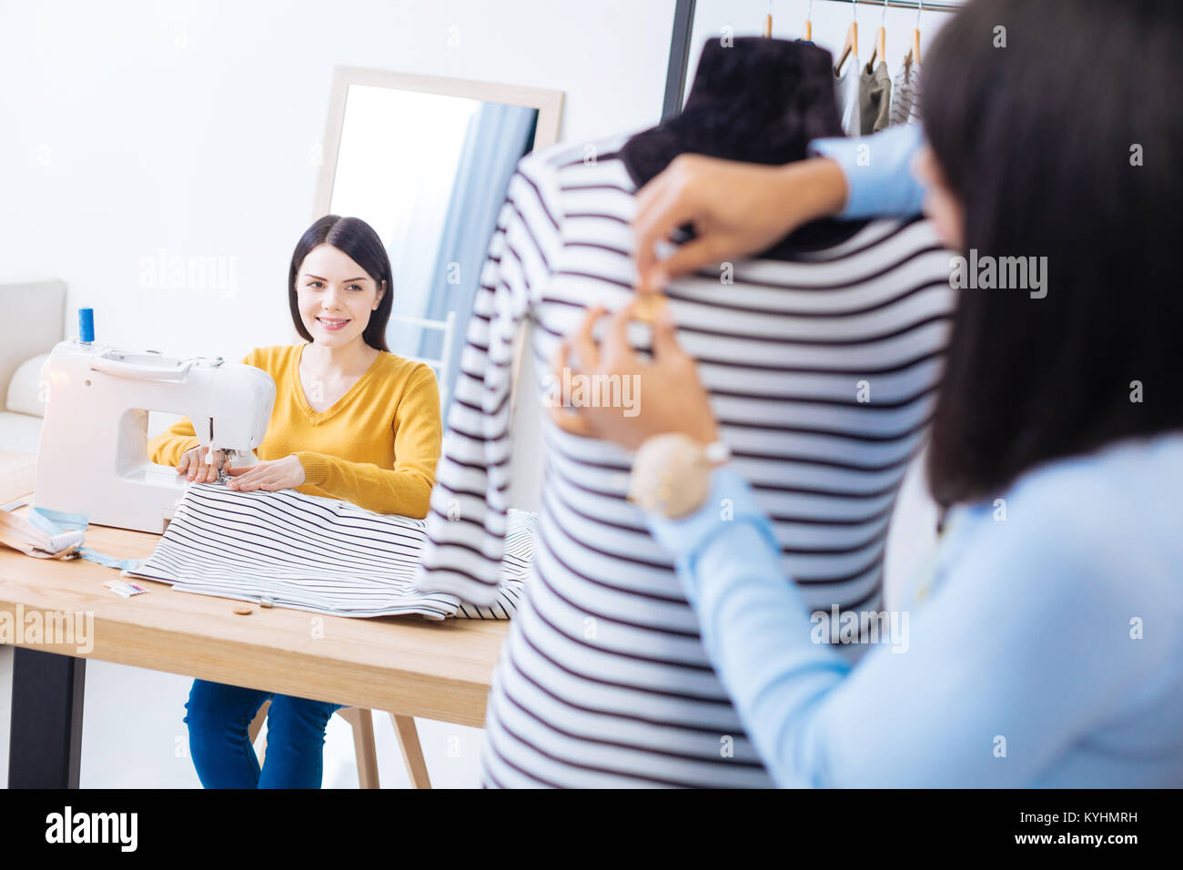 Polite tailor smiling to her colleague while sitting and sewing Stock ...