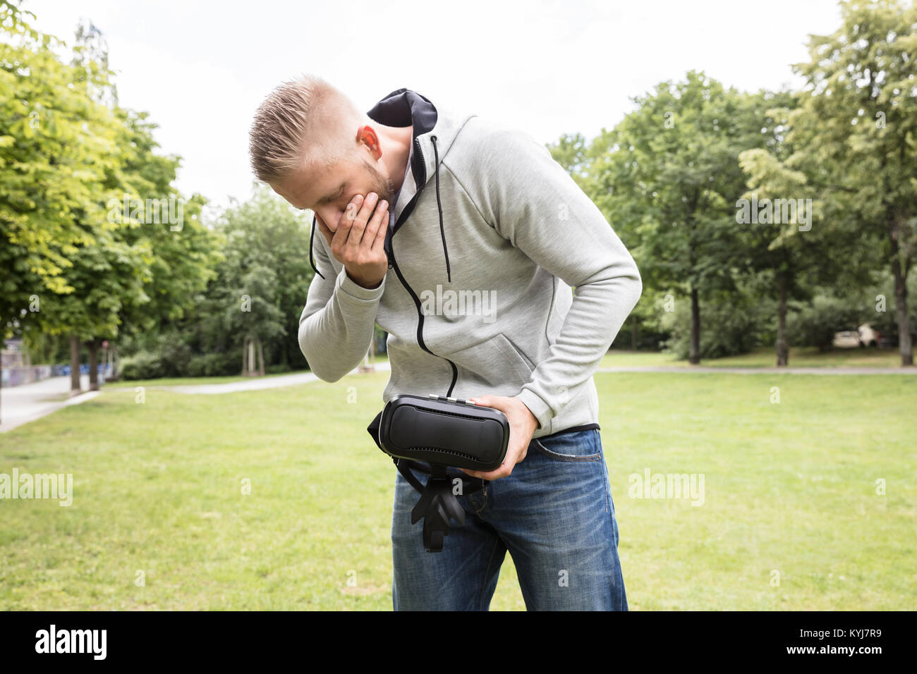 Young Man With Nausea Holding Virtual Reality Headset In The Park Stock