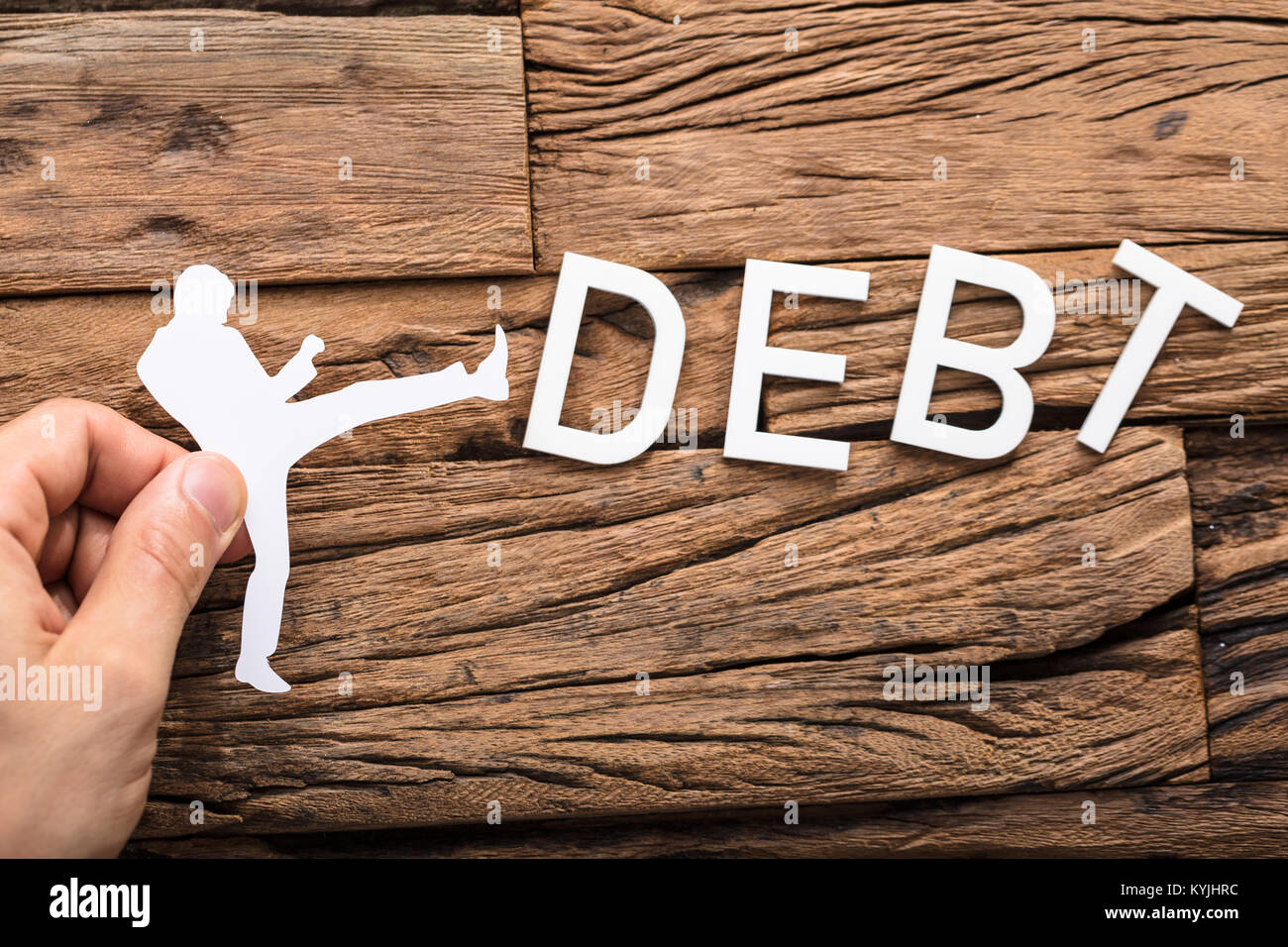 Hand Holding A Paper Cut Out Figure Kicking The Debt Word On Wooden Table Stock Photo