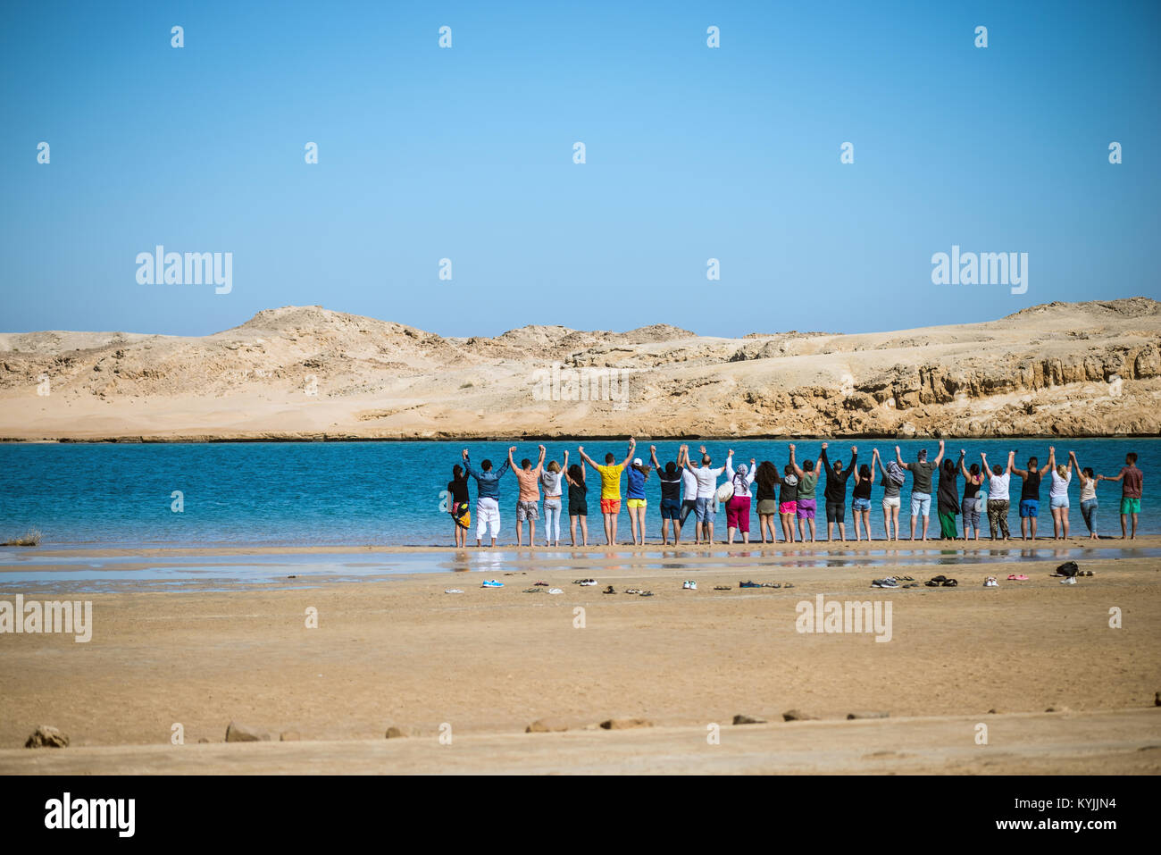 A group of people holding hands each other and standing near a blue lake surrounded by a desert Stock Photo