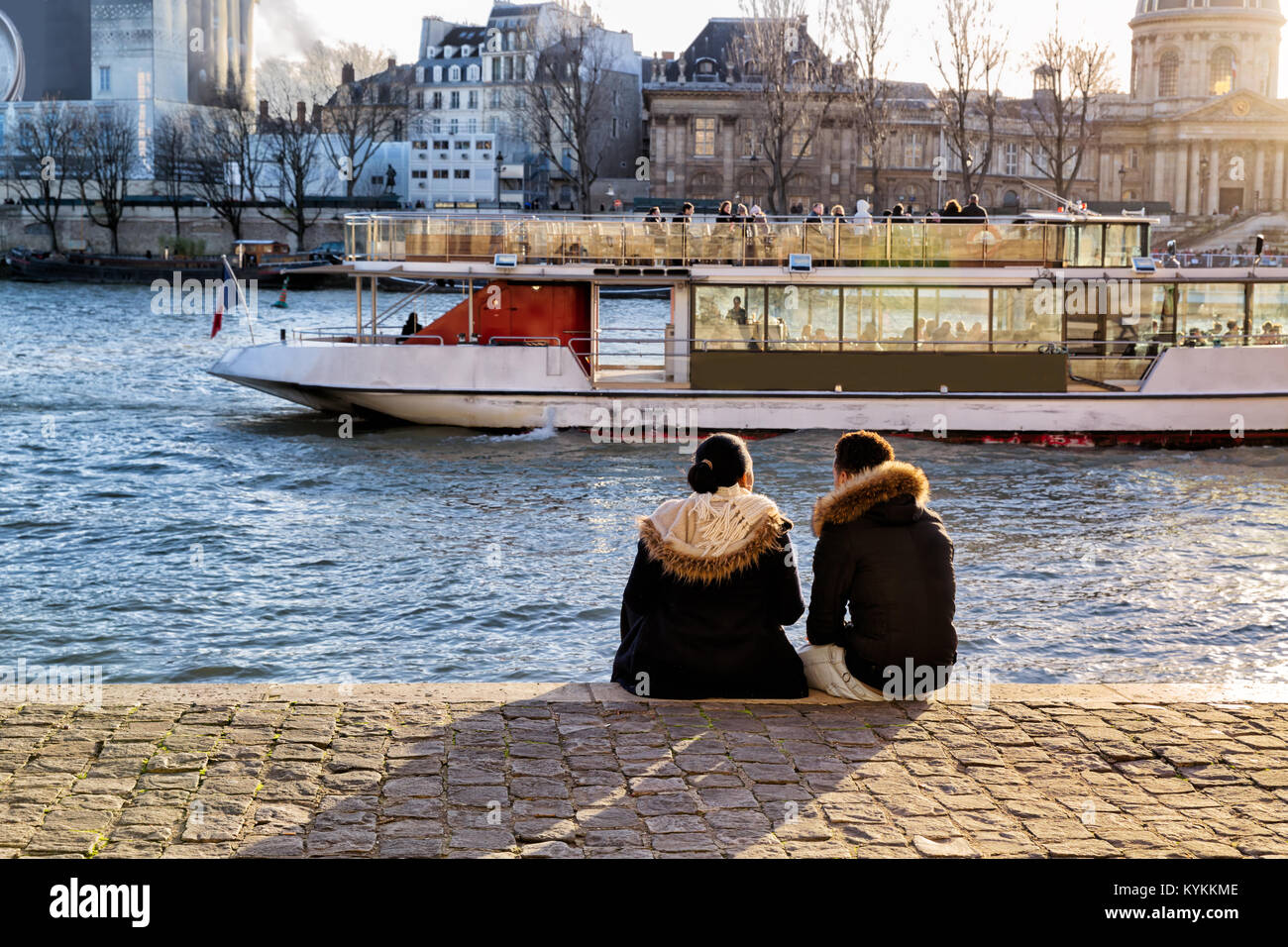 Paris young couple sit on the Seine river bank watching the sunset and a boat going by. They wear fur collars on the cold winter day. Romantic view. Stock Photo