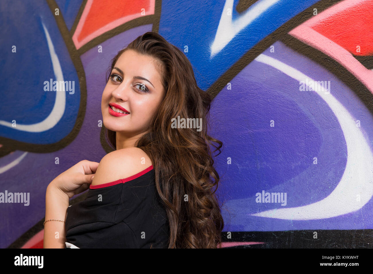 street fashion, portrait near the wall with graffiti. girl brunette chubby lips with red lipstick and curly hair. Stock Photo