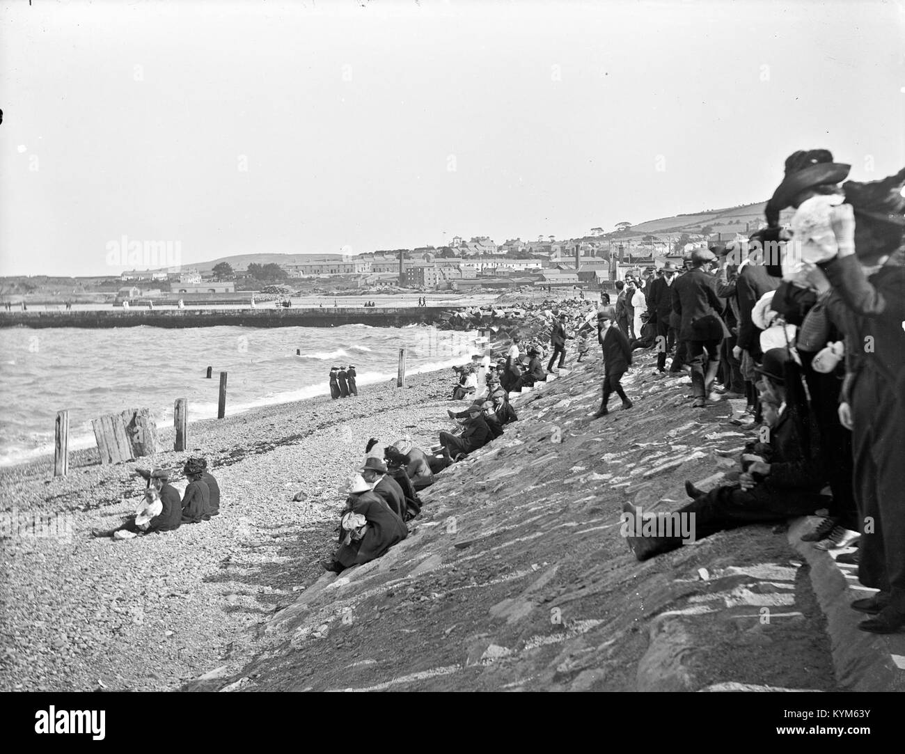 This image, part of the Lawrence Collection, depicts a crowd at a bathing strand in County Wicklow. The photograph captures a vintage seaside scene, featuring a gasometer, pier, and groyne, with a likely regatta in the background. Stock Photo