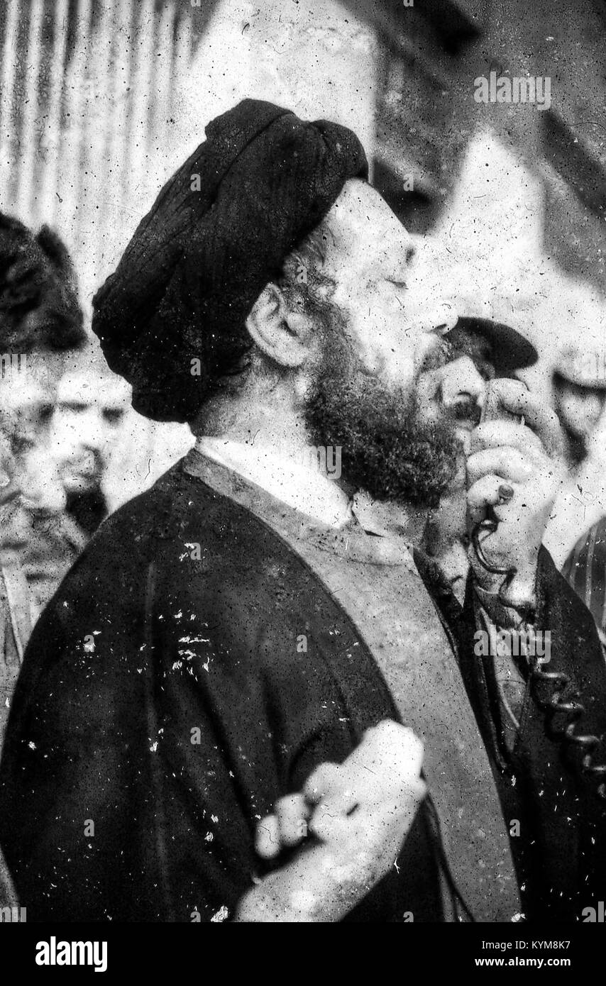 View of a man dressed in traditional clerical robes and black turban, speaking into a loudspeaker microphone at a rally in Iran, March, 1983. Extensive damage on original photo. () Stock Photo