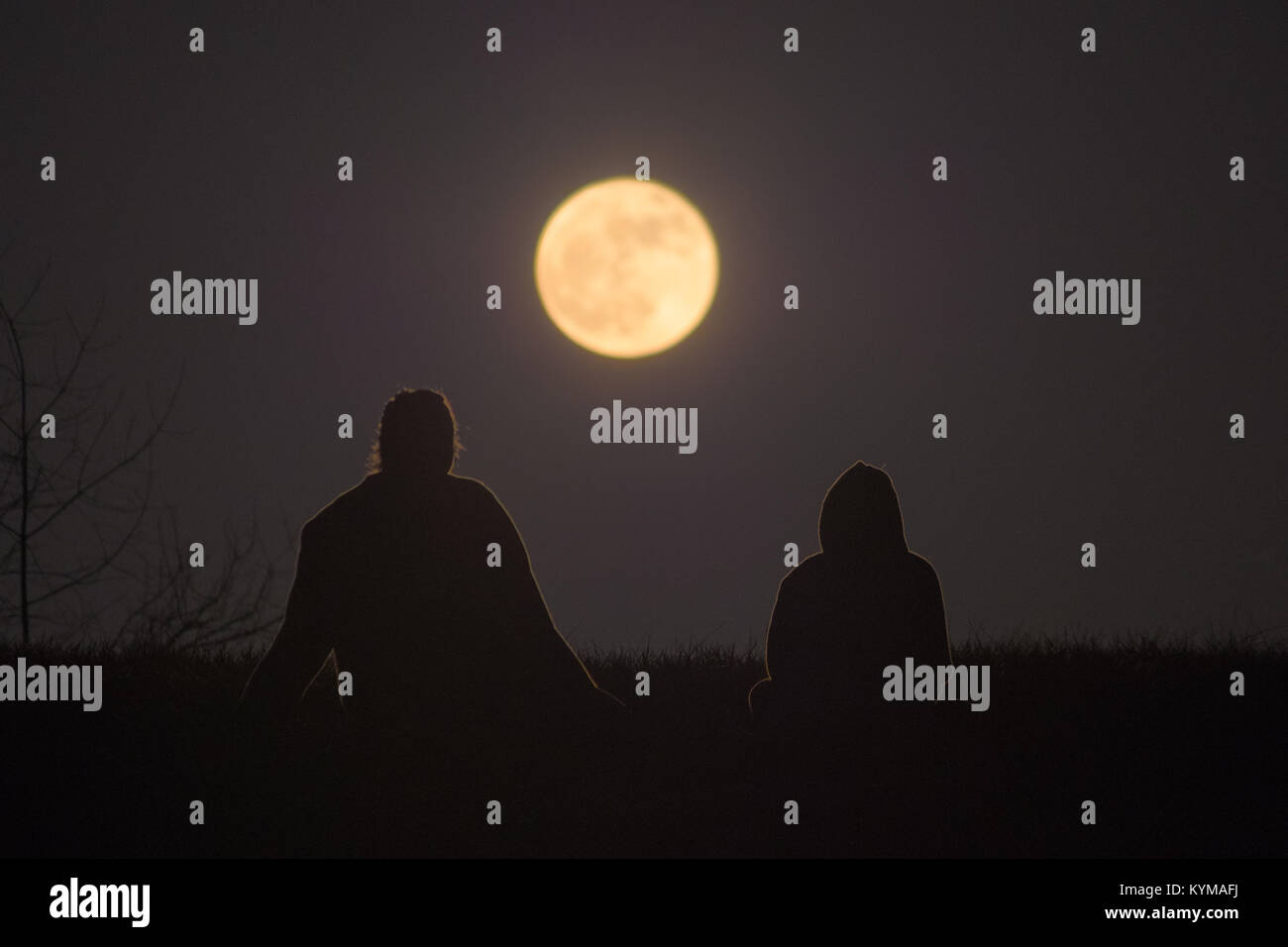 Two women are sitting on the ground against the horizon, the full moon rising between them. Stock Photo