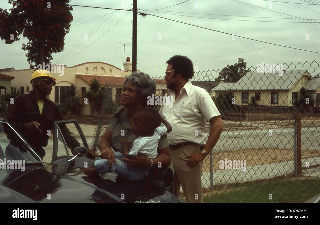 Scene of an African American family outside of their home in Los Angeles, California, 1977. An African American man wearing a yellow hardhat talks with another man as he moves to sit behind the wheel of a 1969 Chevrolet Chevelle. A gray-haired African American woman steadies an infant African American boy sitting on the hood of the car. () Stock Photo