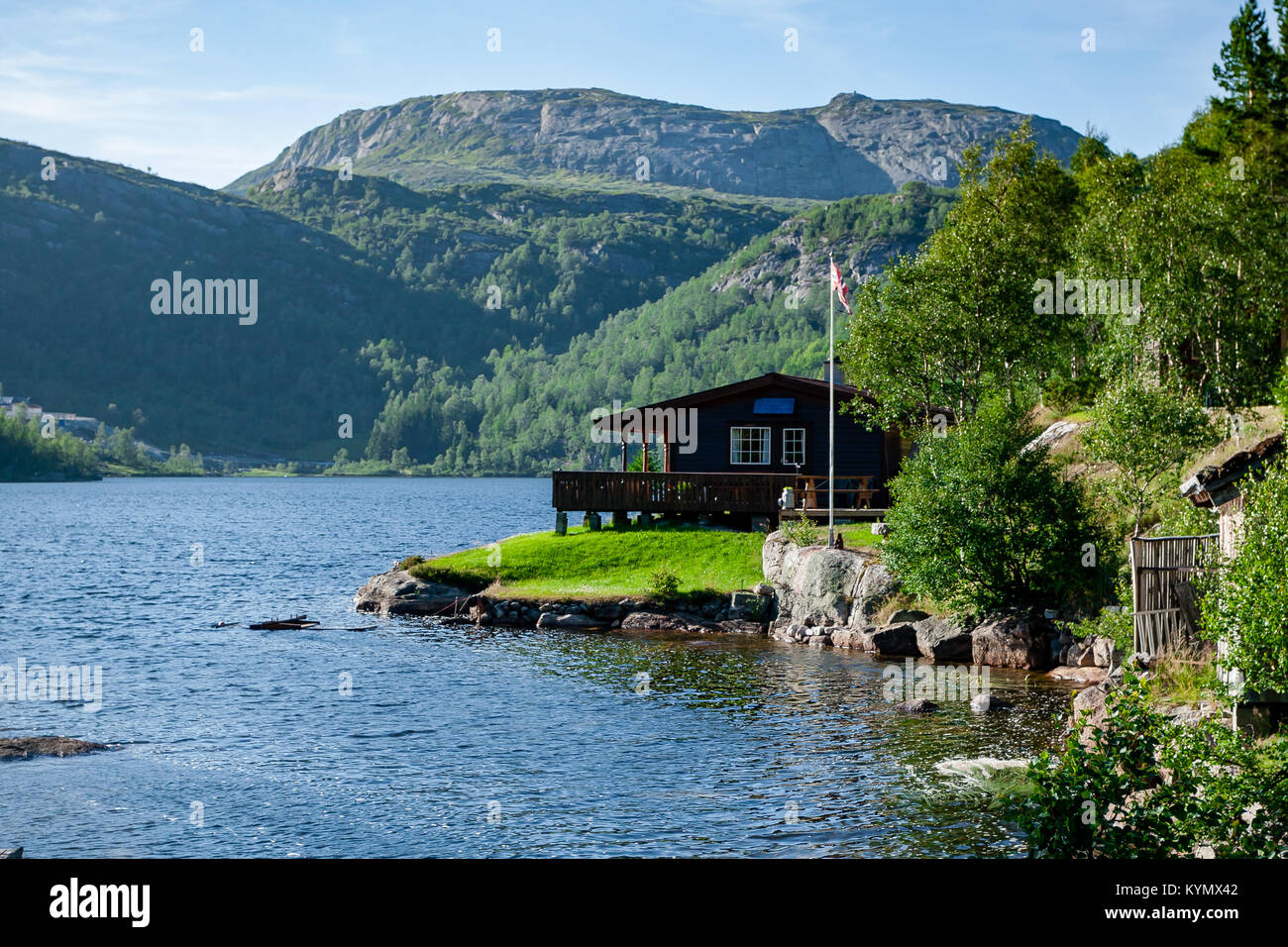 Norwegian wooden house build by the fjord with the scenic mountain view in the background. Norwegian flag. Blue sky. Idyllic location Stock Photo