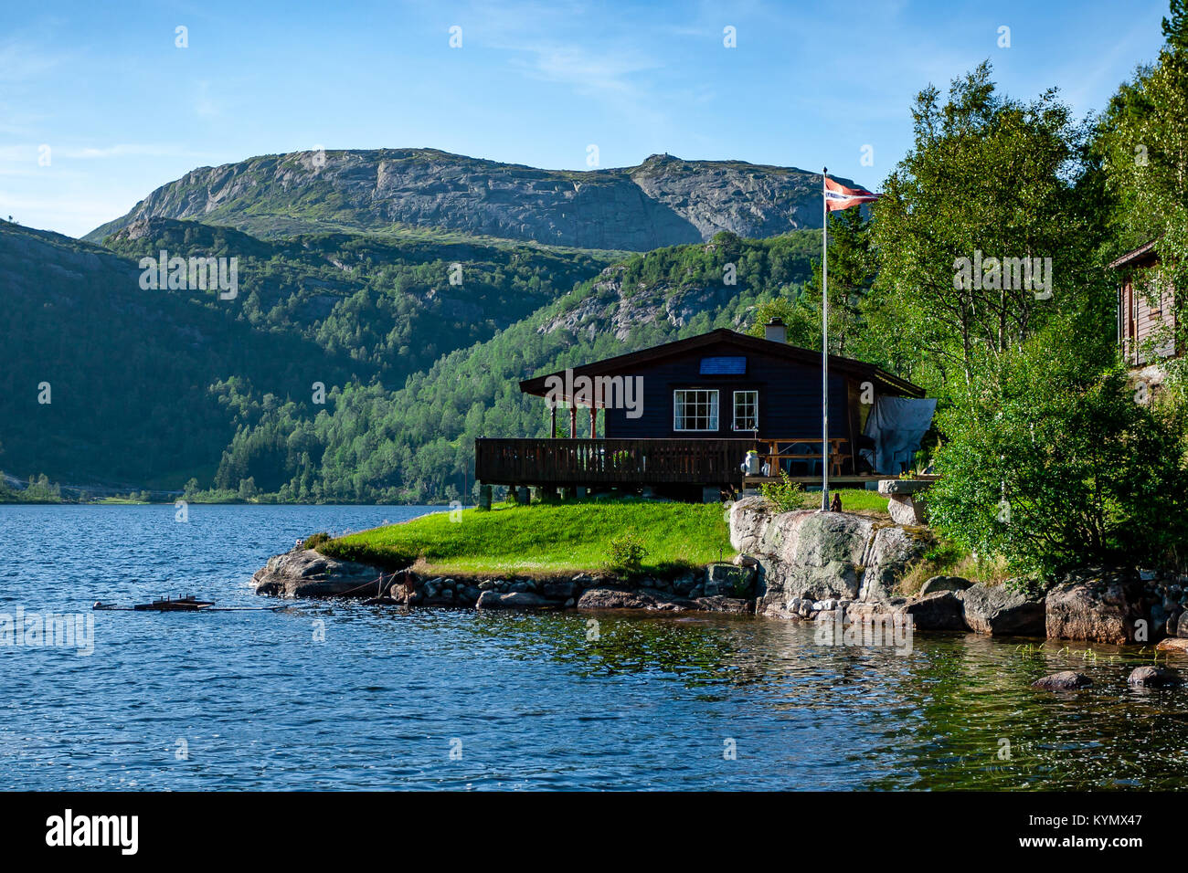 Norwegian wooden house build by the fjord with the scenic mountain view in the background. Norwegian flag. Blue sky. Idyllic location Stock Photo