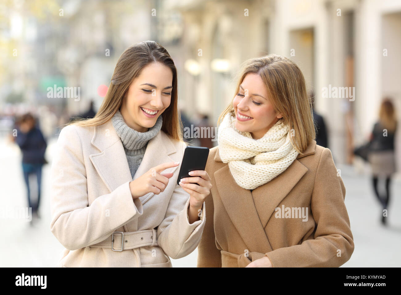 Front view of two friends walking and watching smart phone content in winter on the street Stock Photo