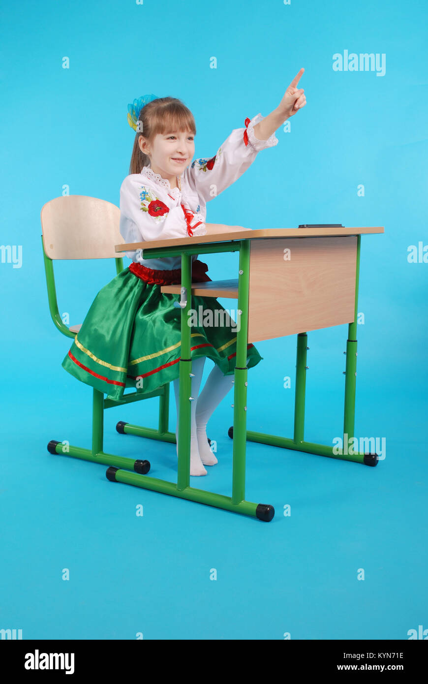 Young girl with light hair in Ukrainian embroidery sitting on school desk isolated on blue backround Stock Photo