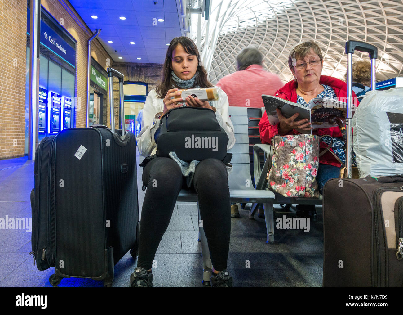 Two women sitting on a bench with luggage, one reading a magazine, the other about to eat a sandwich. Kings Cross train station, London, England, UK. Stock Photo