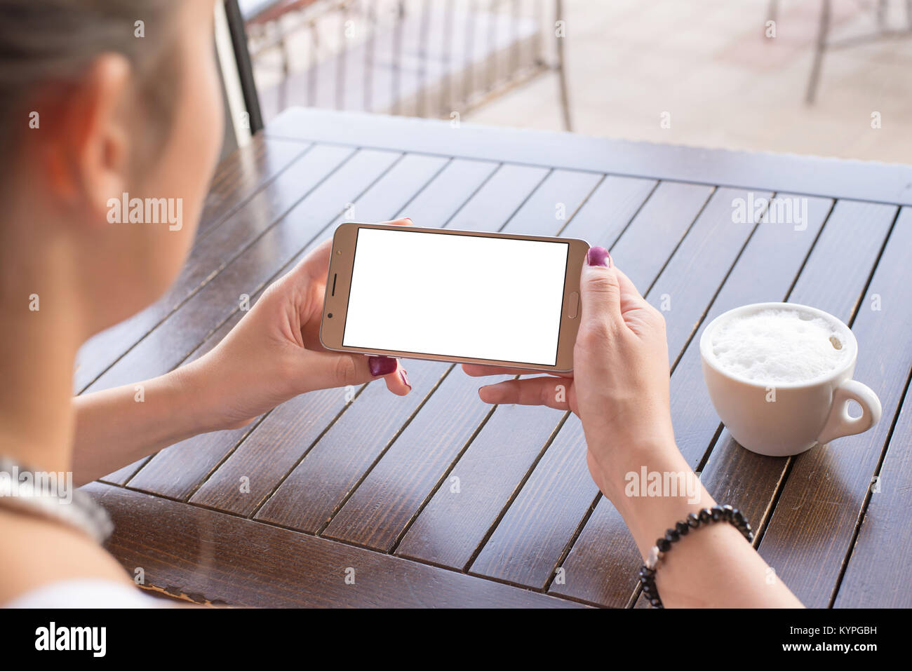 Woman holding mobile phone in horizontal position with isolated screen for mockup. Coffee cup and wooden desk in background. Stock Photo
