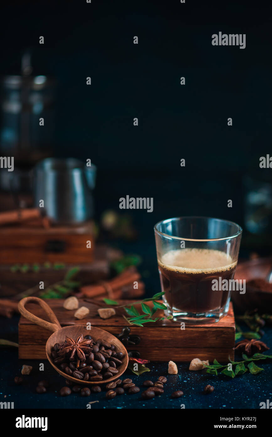 Espresso shot with foam on a wooden box with coffee beans, arabica leaves, cinnamon and spices on dark background. Brewing coffee in French press. Stock Photo