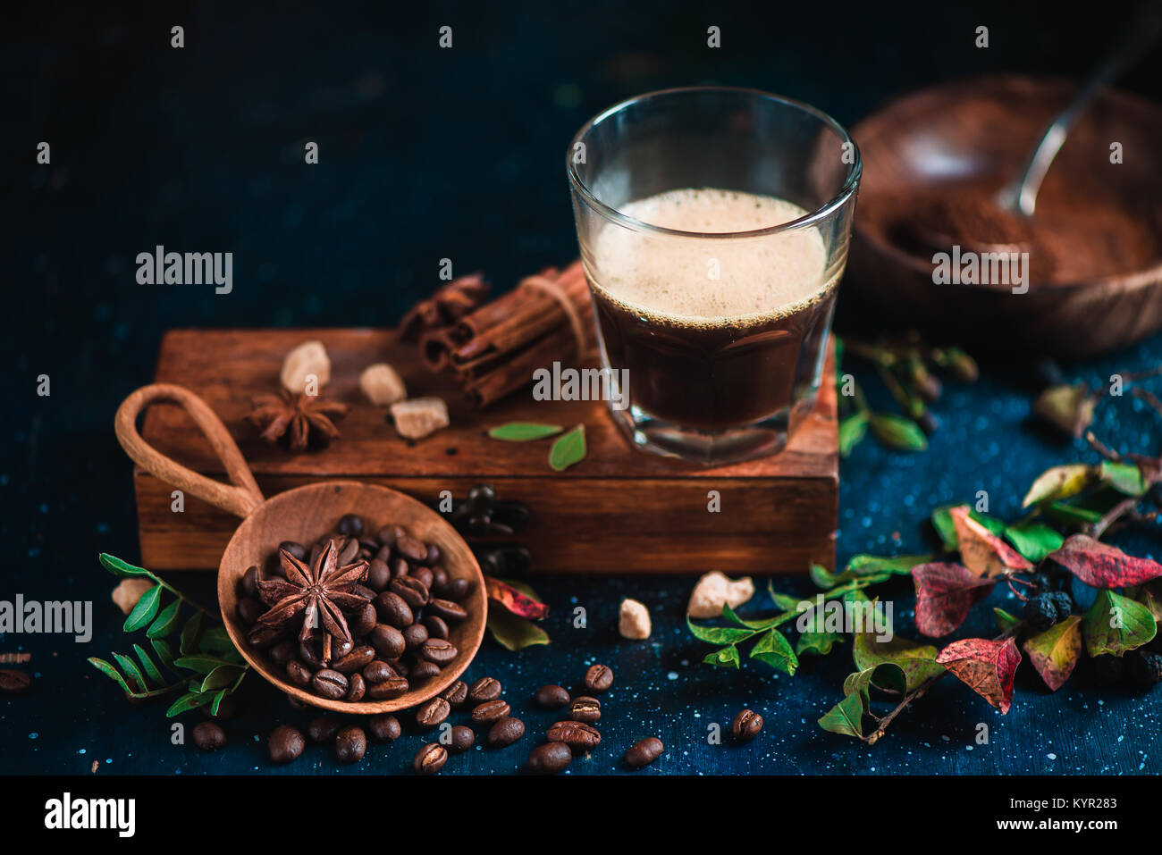 Espresso shot with foam on a wooden box with coffee beans, arabica leaves, cinnamon and spices on dark background. Brewing coffee still life. Stock Photo