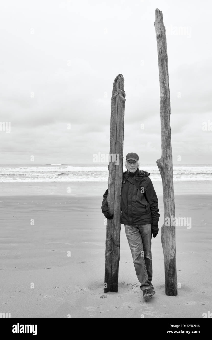 Portrait of senior man on the beach standing with driftwood posts in the sand. Ocean in the background.  Black and white monochrome. Copy space. Stock Photo
