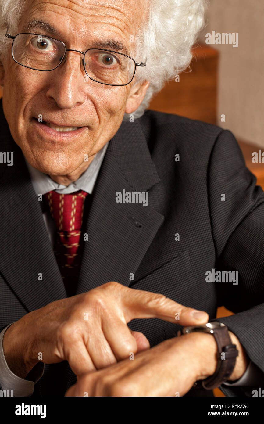 Impatient old man pointing at his watch. Looking at camera with a look of exasperation on his face. Wearing a business suit. Close up Stock Photo