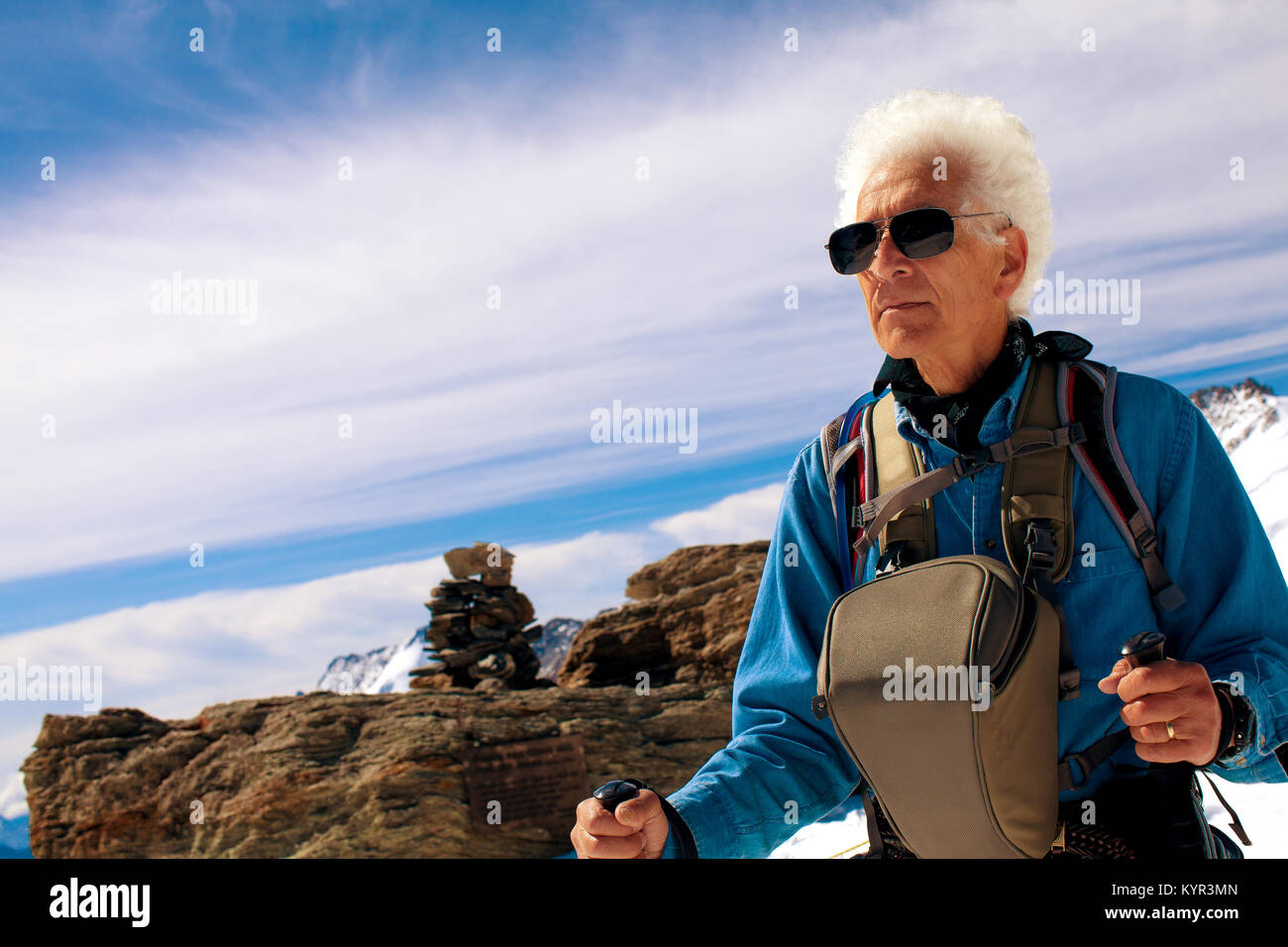 Senior man hiking in the mountains on adventure travel trip to the Swiss Alps. Wearing sunglasses, camera bag, trekking poles. Copy space. Stock Photo