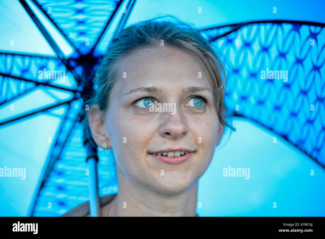 A blonde girl with blue eyes smiling under a blue umbrella Stock Photo