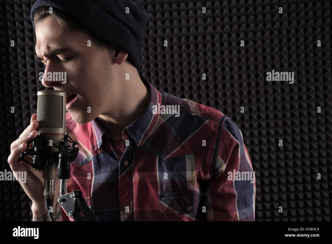 Close-up portrait of emotional young guy singing with his eyes closed and his brow in a knit in a cap standing in front of the microphone stand holdin Stock Photo