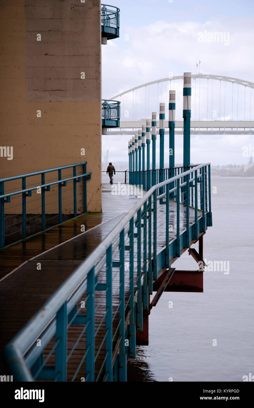 A man walks along the wooden deck of the Willamette River in Portland with a fence and lined lanterns in the rainy Portland weather Stock Photo