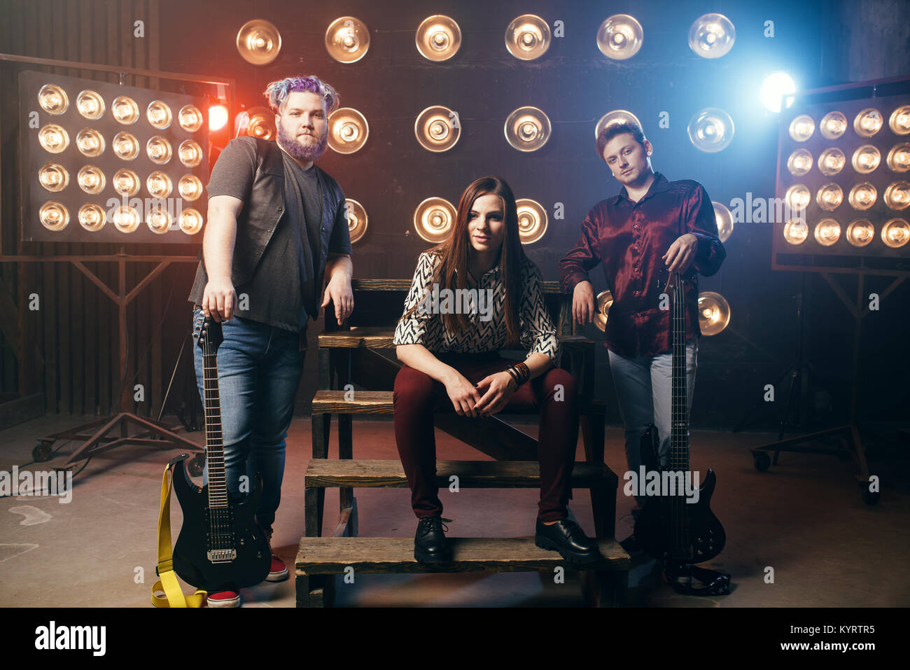 Musical group poses on the stage with lights in night club, vintage style. Guitarists and drummer, rock band concert, music show and entertainment Stock Photo