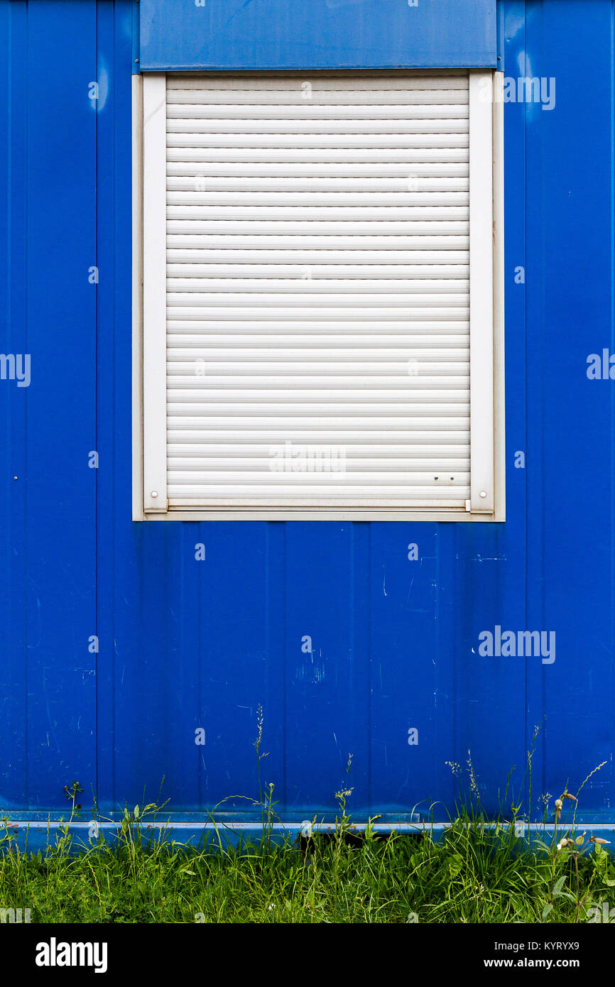 Window with closed blinds on a blue tin wall with green grass stripe Stock Photo