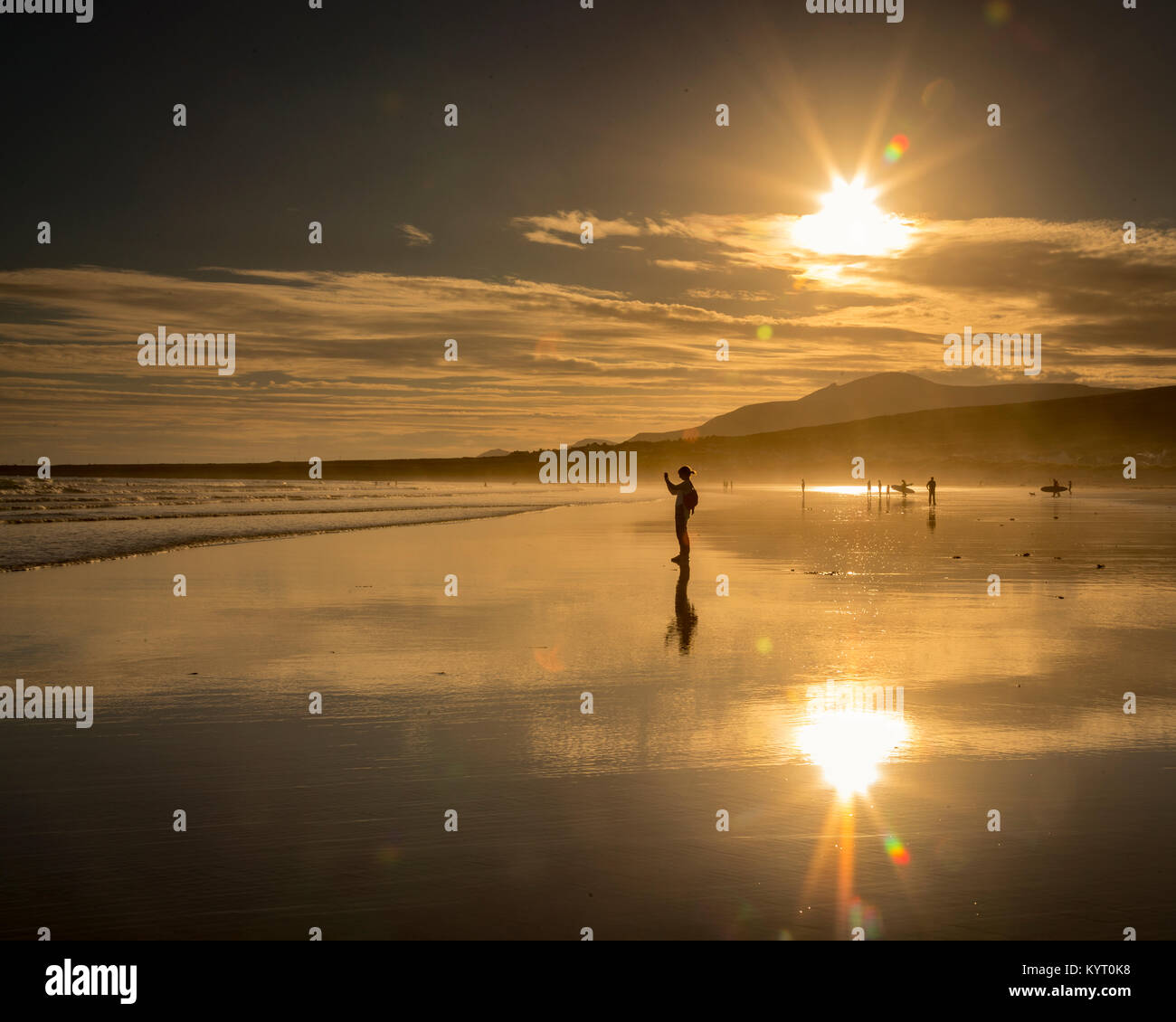 Girl in silhouette taking a photo while standing on reflective wet sand on Keel beach on the west coast of Ireland with the sun shining above. Stock Photo