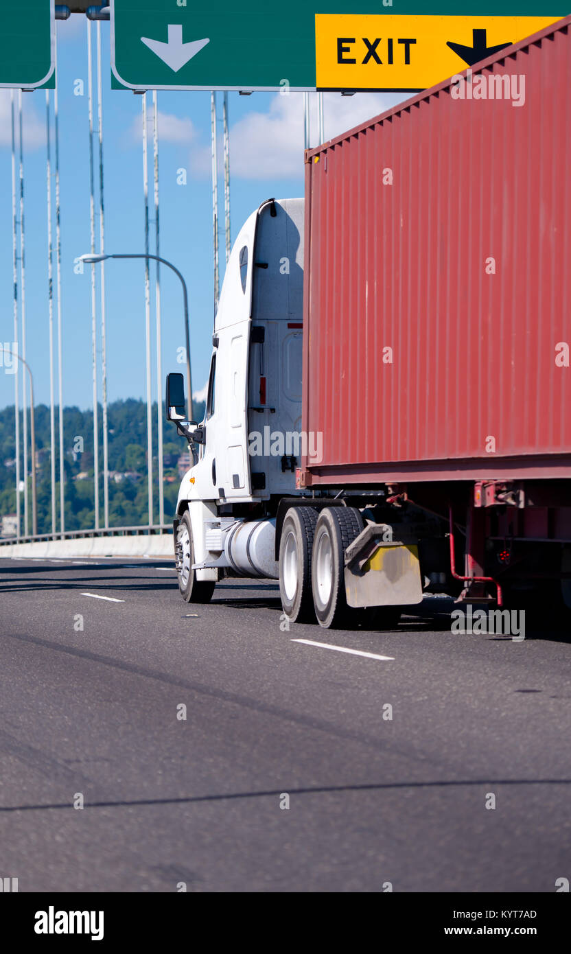A large professional white modern big rig semi truck with a flat bed trailer, on which a ribbed long container is fixed, moves along an roadway Stock Photo