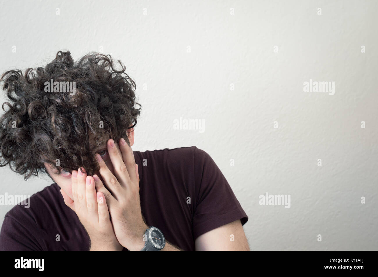 Portrait of a young, Caucasian, brunet, curly haired man covering his face with hands on a white background. Stock Photo