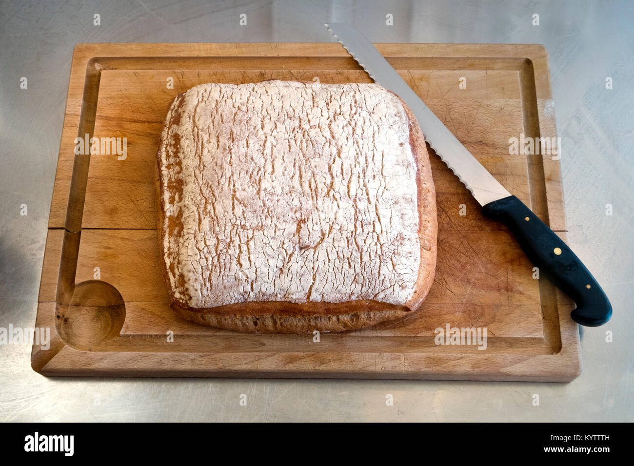 A rustic artisan style French bread loaf waits on a chopping board on a stainless steel kitchen work surface Stock Photo