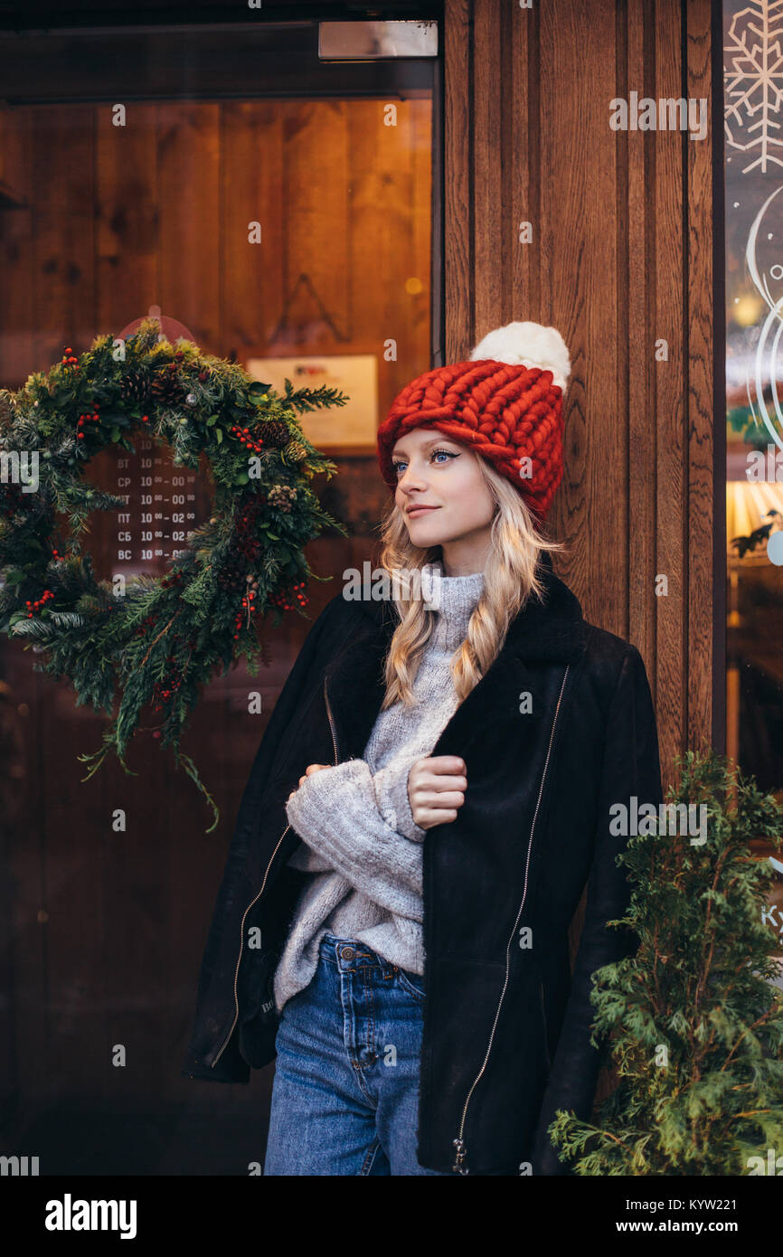 Young blonde woman in red knitted hat with white pompon, grey sweater, black jacket and blue jeans make posing with Christmac cafe window on the backg Stock Photo