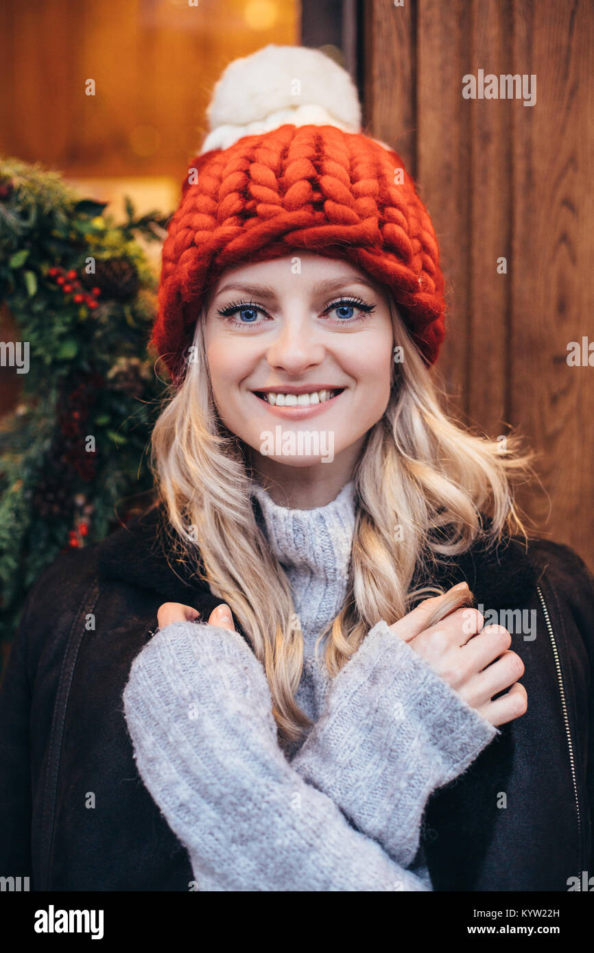Young blue eyed blonde woman in red knitted hat with white pompon, grey sweater and black jacket make posing with Christmac cafe window on the backgro Stock Photo