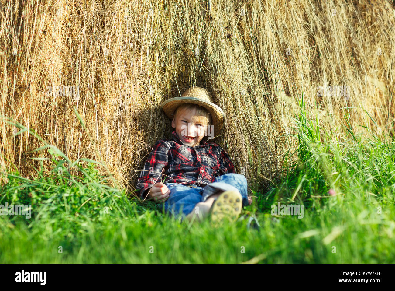 Happy boy in straw hat sitting in haystack, outdoors, looks camera. Smiling child relax haystack on green grass under the hot sun. Young man spends ho Stock Photo