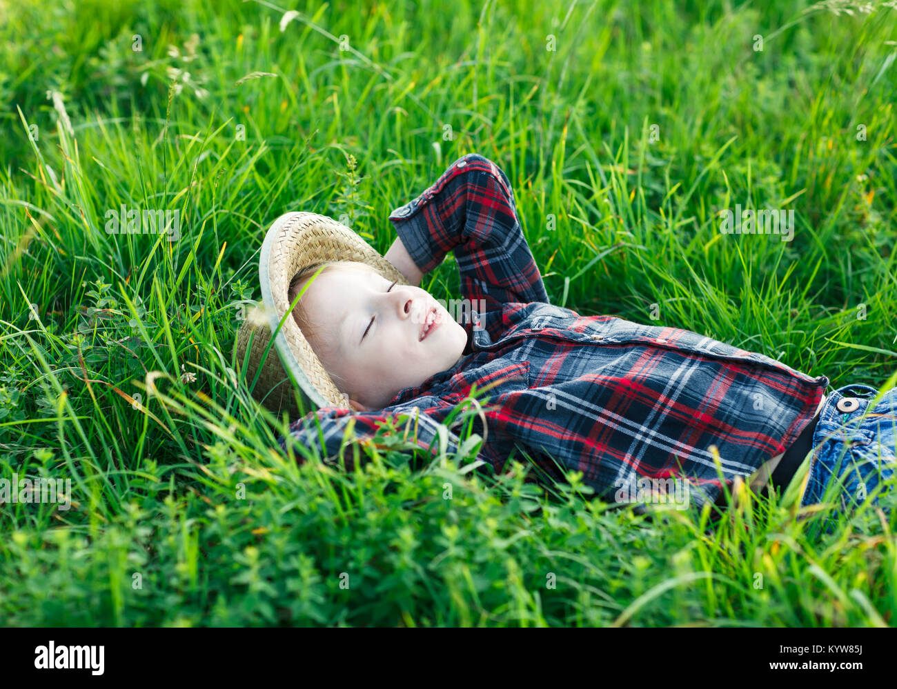 Handsome little boy lying on grass on back, hands behind head. Little dreamer lies in meadow with hat, eyes closed. Child sleeping and smiling in natu Stock Photo