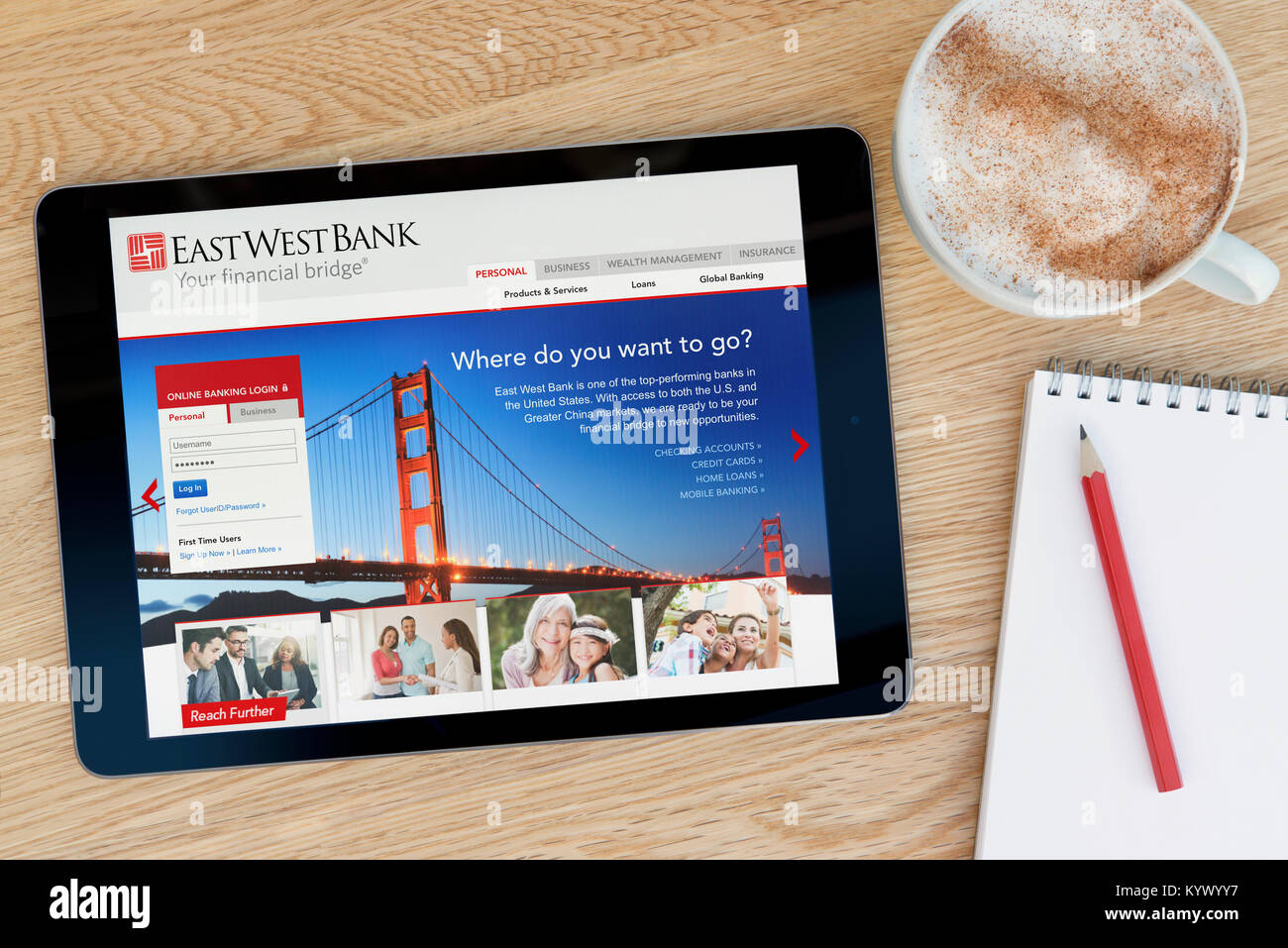 The East West Bank website on an iPad tablet, on a wooden table beside a notepad, pencil and cup of coffee (Editorial only) Stock Photo