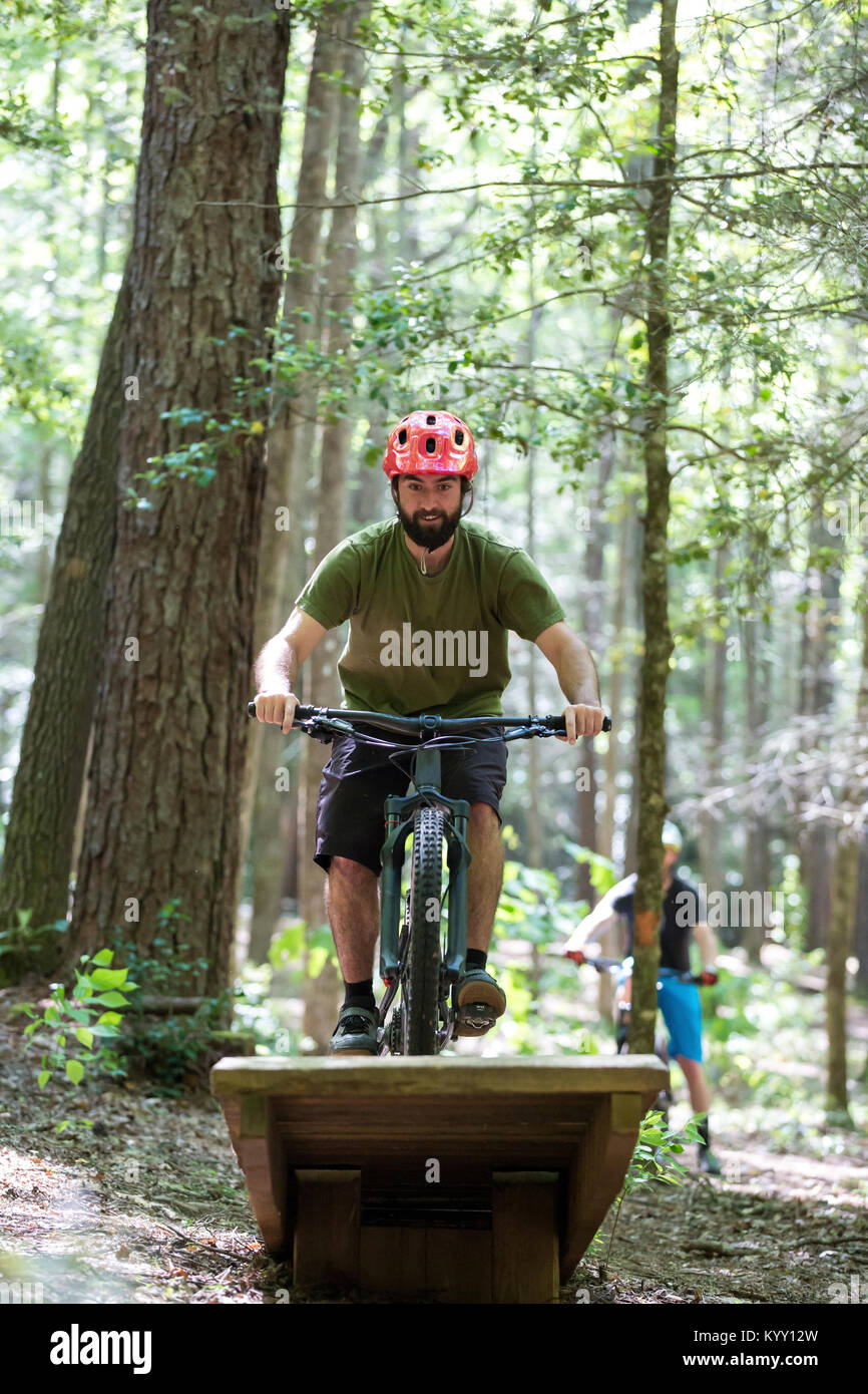 Hiker doing stunt while riding mountain bike with friend in background at forest Stock Photo
