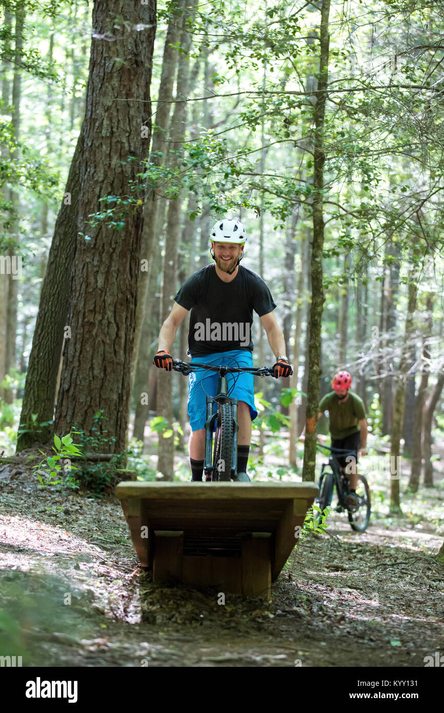 Happy hiker doing stunt while riding mountain bike with friend in background at forest Stock Photo