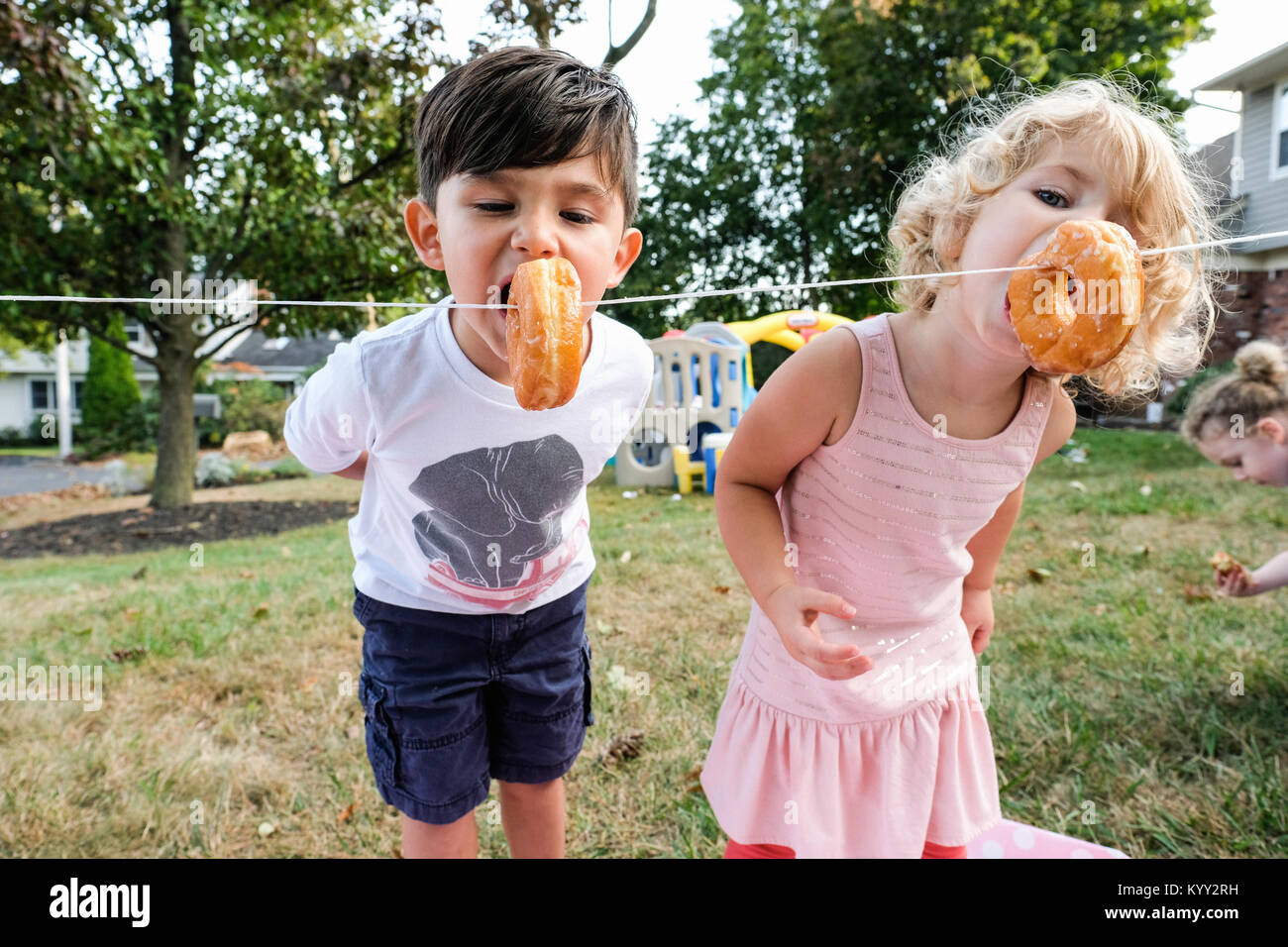 Siblings eating donuts hanging on rope in yard during celebration Stock Photo