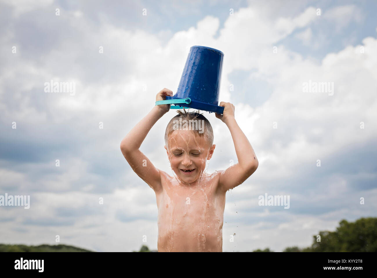 Shirtless boy pouring bucket of water over head against cloudy sky