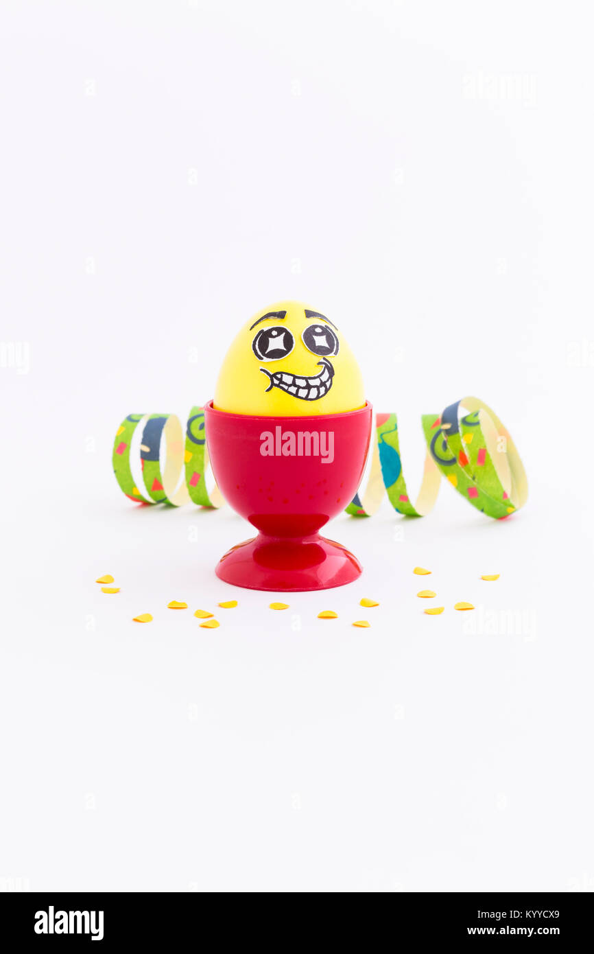 Yellow painted Easter egg with funny cartoon style face in a red plastic egg cup, colorful paper streamer and confetti on white background Stock Photo