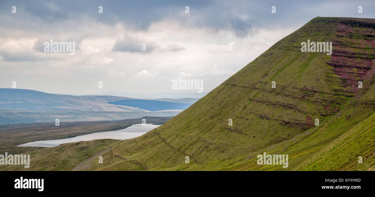 A lone walker stands on the summit of Fan Brycheiniog overlooking Llyn y Fan Fawr galcial lake in the Brecon Beacons of South Wales UK Stock Photo