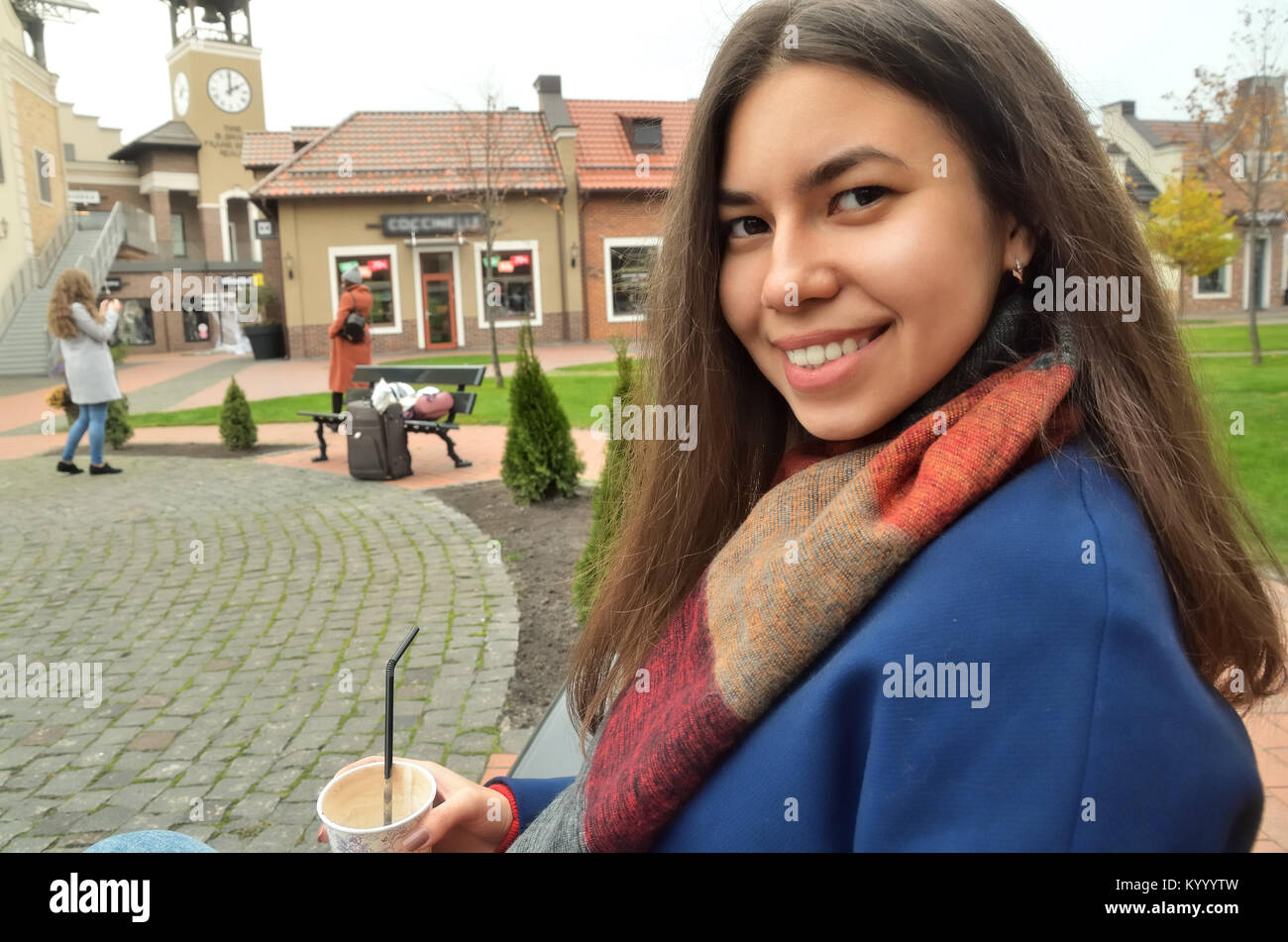 Portrait of a smiling Asian young woman sitting on a bench with coffee. Around the boutiques, shops, trees and greens. Horizontal photo Stock Photo