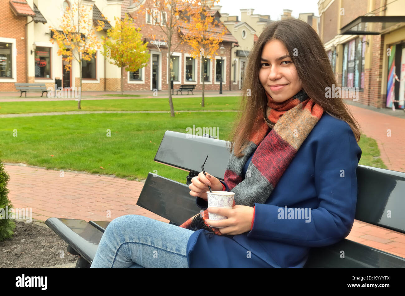 Asian girl sits on a bench outdoor with a cup of coffee in her hands. Smiles and looks at the photographer. Dressed in a coat and a scarf. Hair split Stock Photo