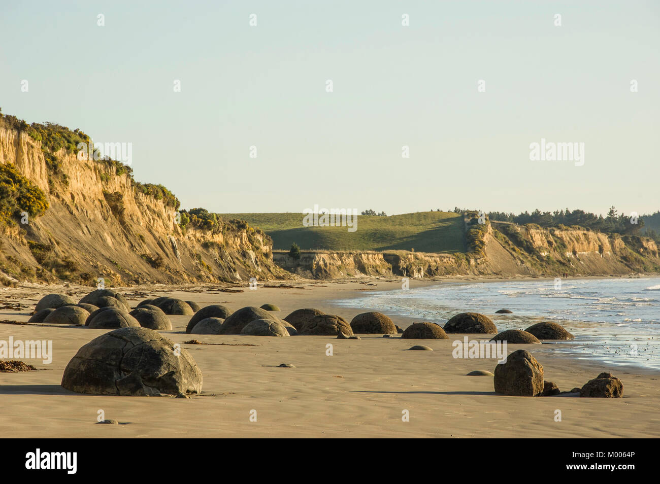 Large stones called Moeraki Boulders on Koekohe beach with sunrise reflecting off cliffs, 2 people walking, East Coast, South Island, New Zealand Stock Photo