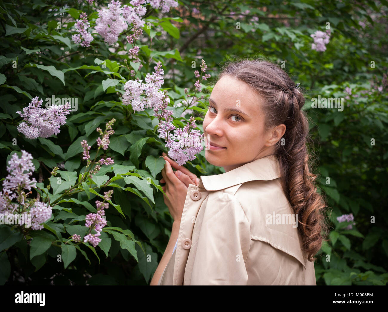 Young girl is standing near a blooming lilac bush and looking at camera. Stock Photo