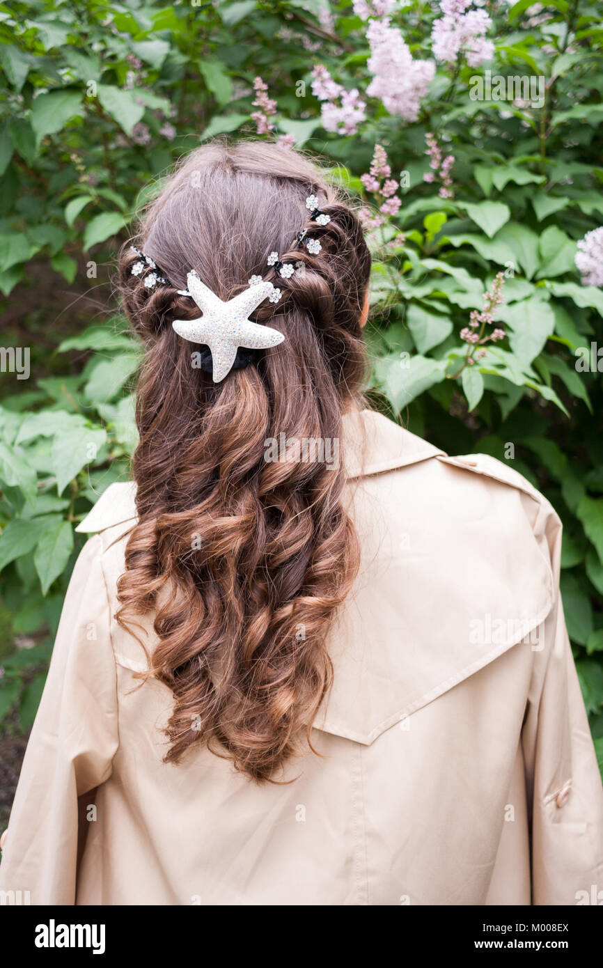 Young girl with curly hair is standing near blooming lilac bush and looking at it. Rear View. Stock Photo