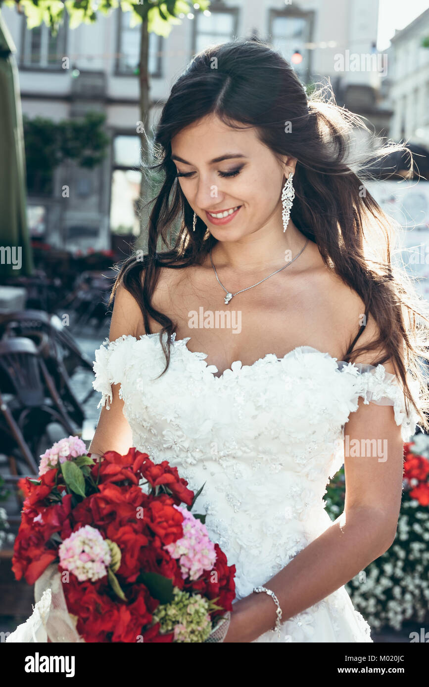 Outdoor portrait of the adorable brunetter bride with pretty smile is holding the wedding bouquet of red and pink flowers. Stock Photo