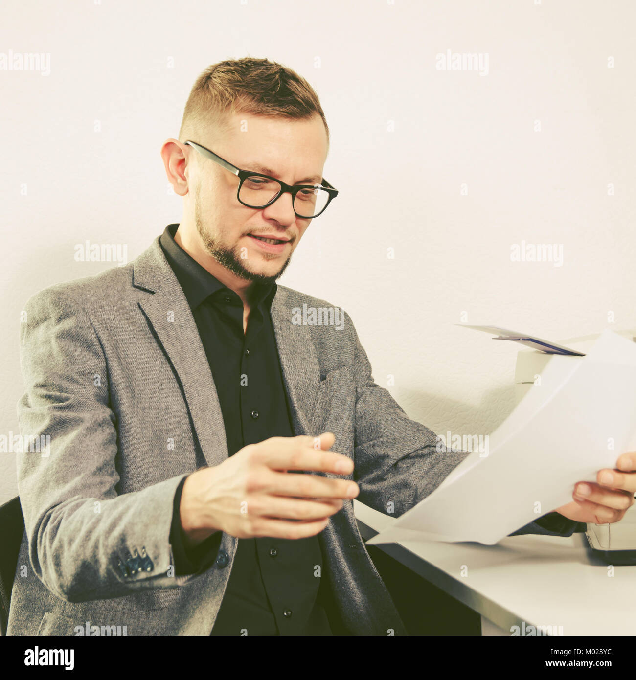 man with glasses and jacket smiles studying documents at table with laptop and printer at office. square view. Stock Photo