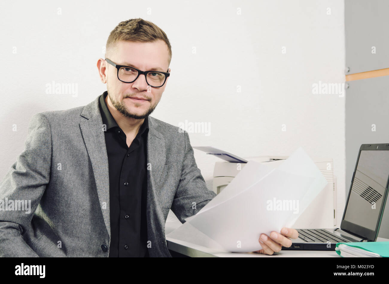 a man in glasses and a jacket with  the documents in his hand smiling looking at lens at the table with a laptop and a printer in the office Stock Photo